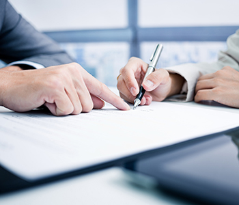 Close-up of two individuals pointing at and signing a document on a desk.